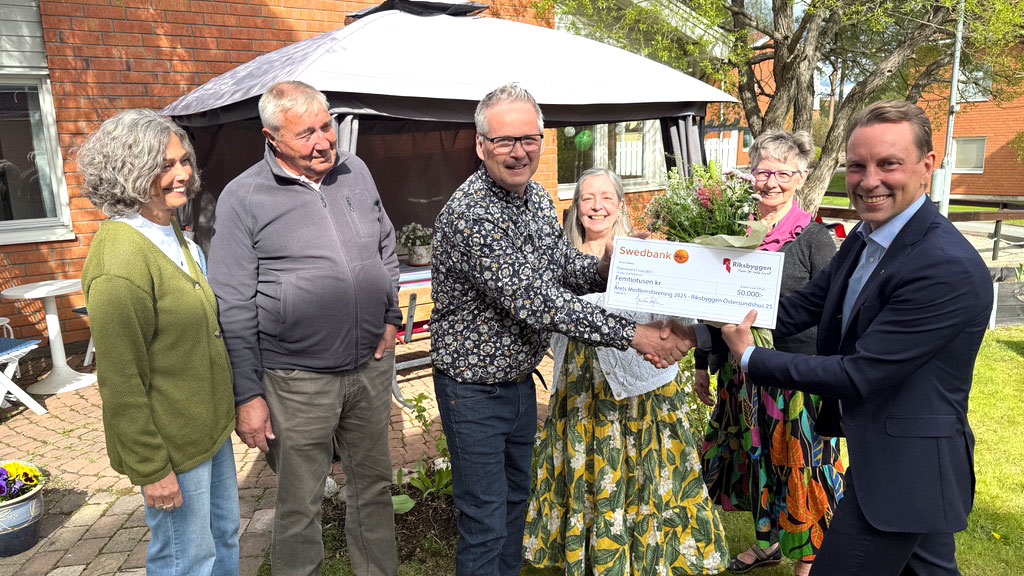 En grupp människor står utomhus och håller en stor check tillsammans, en man i kostym skakar hand med en man i mönstrad skjorta