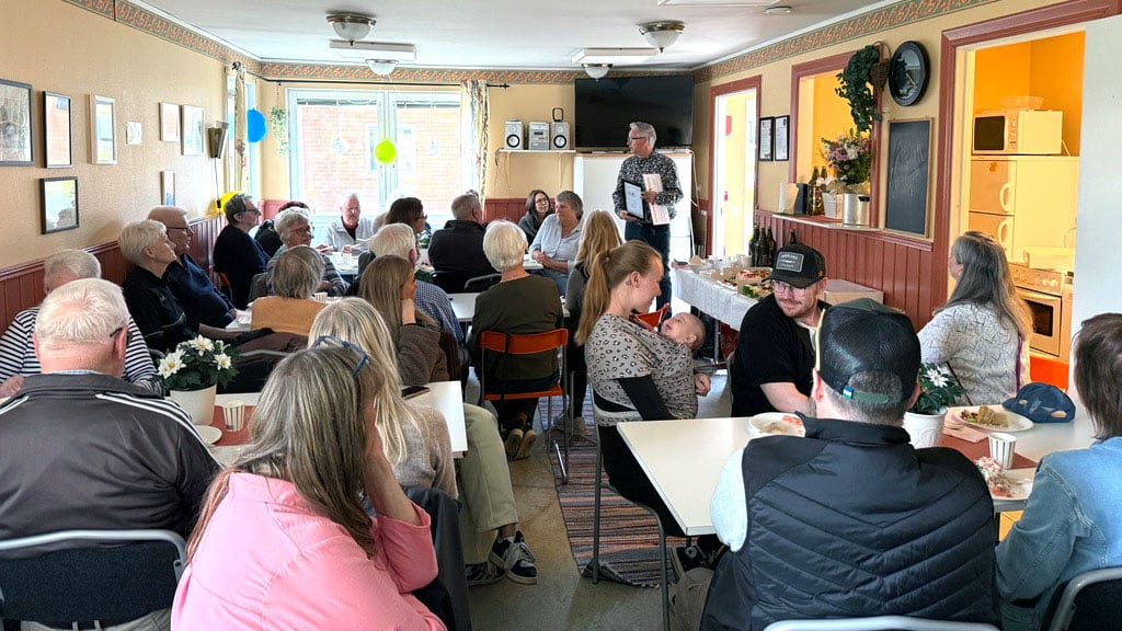 Människor samlade i en möteslokal med dekor och dukade bord, en person står upp och talar