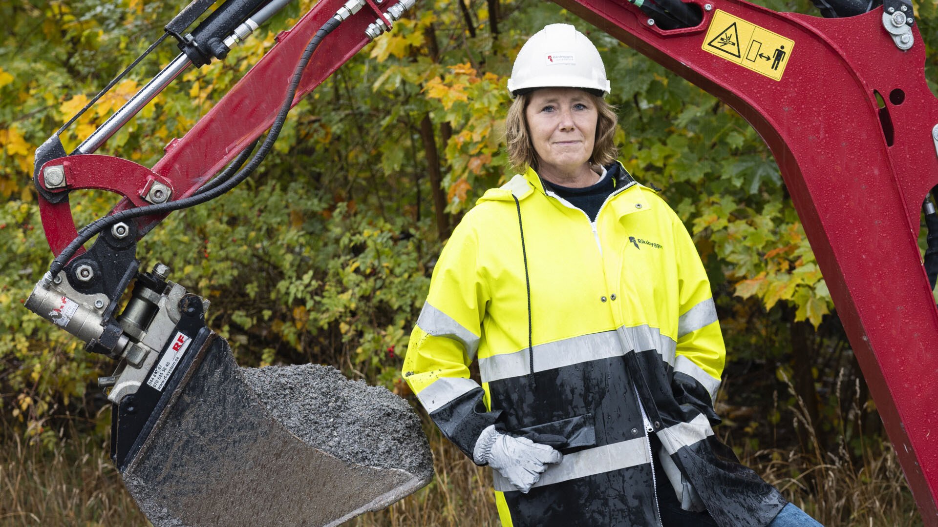 Kvinna i varselkläder och hjälm står vid en grävmaskin framför höstfärgade träd