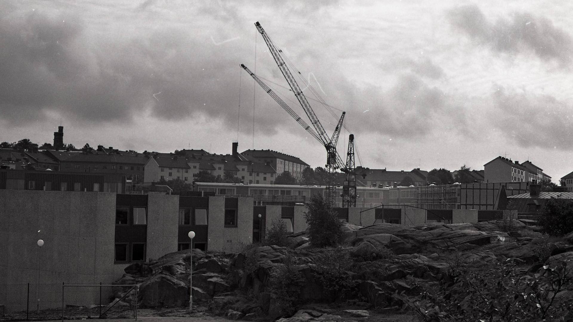 Svartvitt foto av en byggarbetsplats med en stor lyftkran och byggnader i bakgrunden under en molnig himmel