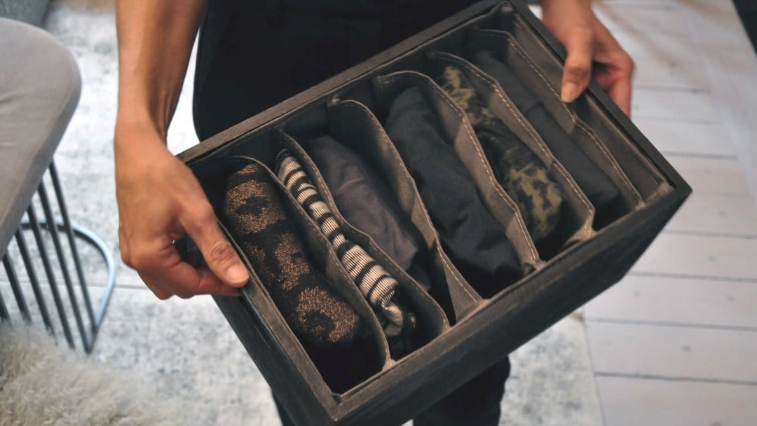 En person håller i en låda med flera fack fyllda med ihoprullade kläder i olika mönster och färger